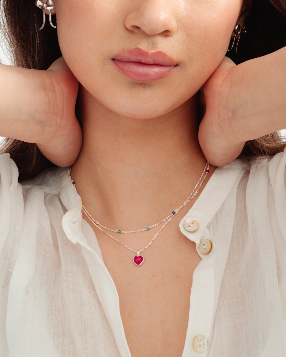 Close-up of a model wearing the silver La La Love Necklace featuring a pink heart pendant, layered with the Santa Monica Necklace with colourful beads. She is also wearing the silver bow-shaped Rosie Earrings.