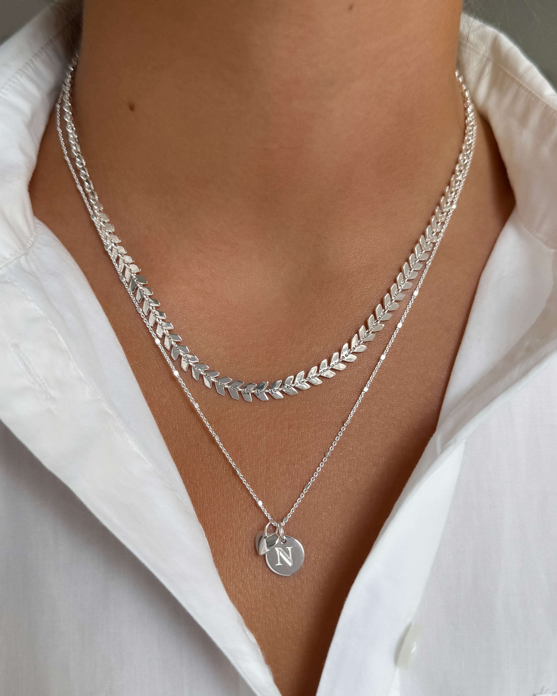 A model wears the silver Layers Olivia Necklace, featuring a delicate leaf pattern, layered with a second fine chain necklace that has a small heart charm and a circular 'N' initial pendant.