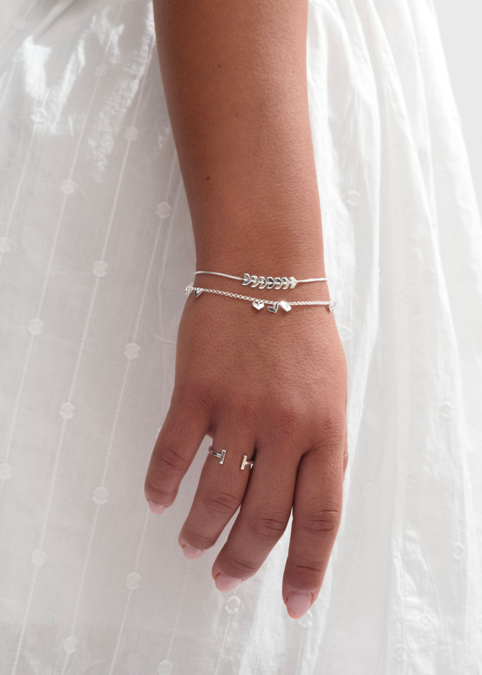 A model's wrist and hand adorned with a stack of silver jewelry: the Layers Simone Bracelet with its leaf-like design, the Layers Bianca Bracelet with small heart charms, and the open Strict Bar Ring.