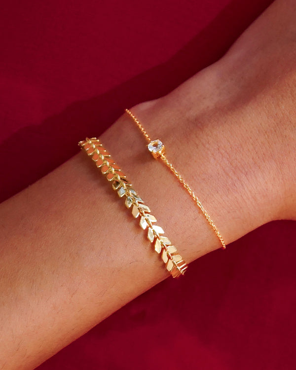 A close-up of a model's wrist wearing the Layers Olivia Bracelet in gold, featuring its signature leaf-like pattern, layered with the delicate Minimalistica Solo Nova Bracelet, a fine gold chain with a single crystal.