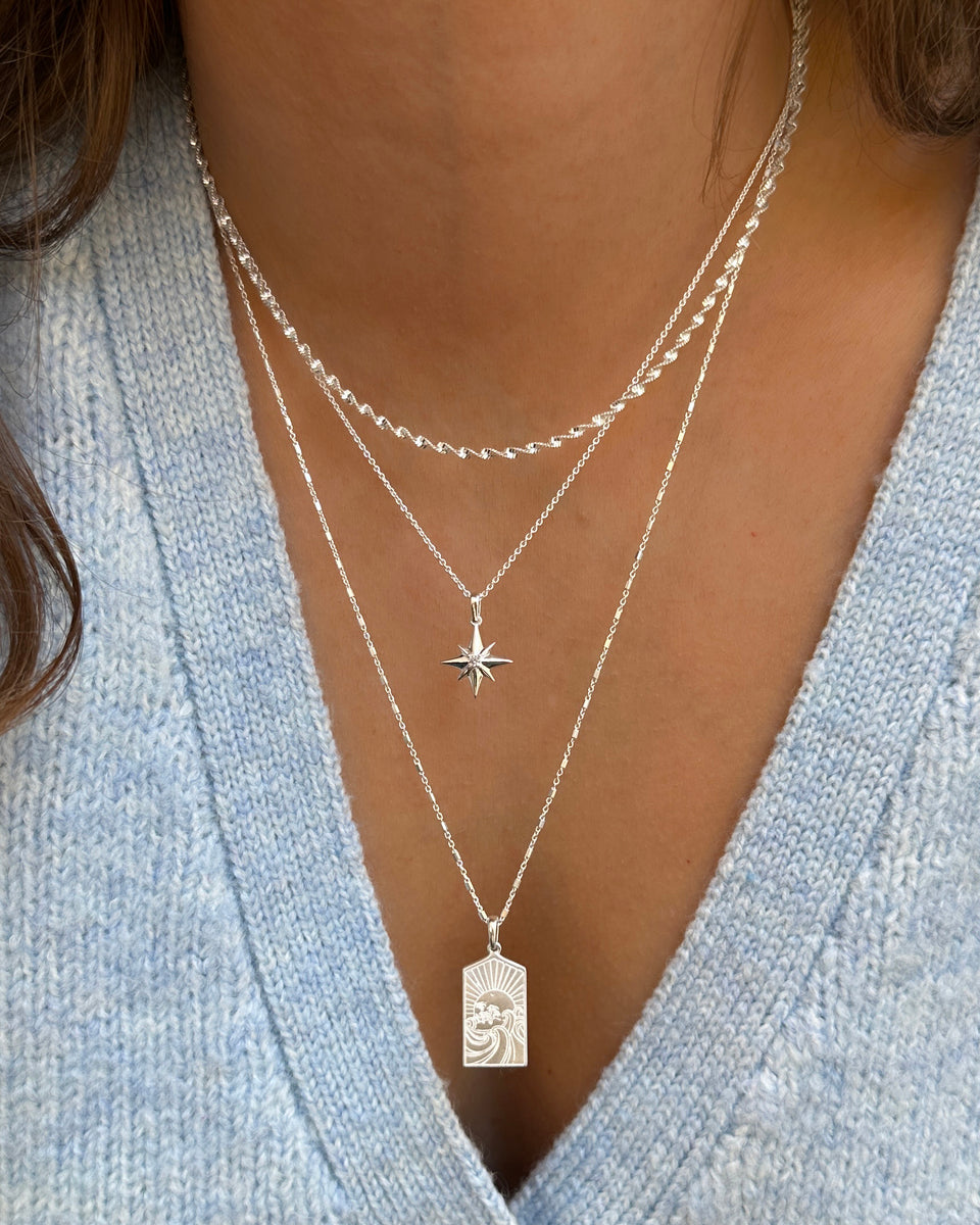 A close-up of a model wearing three layered silver necklaces: the Lucky Coin Keep Dreaming Necklace with its rectangular paradise pendant, the North Star Big Necklace, and the Herringbone Twisted Necklace.
