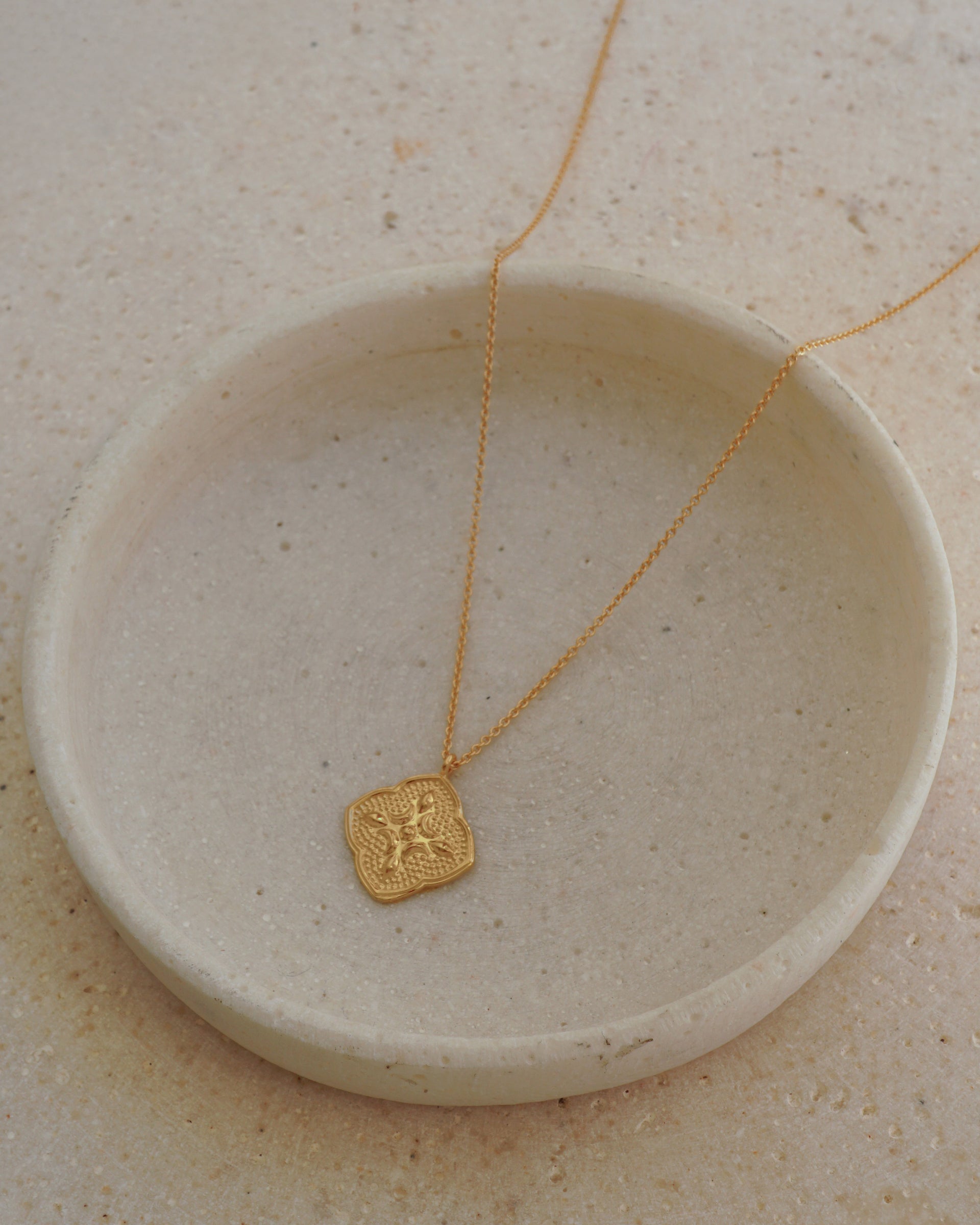 A product shot of the Luna Necklace in gold, featuring a delicate chain and an intricate, quatrefoil-shaped pendant with a textured, embossed design.
