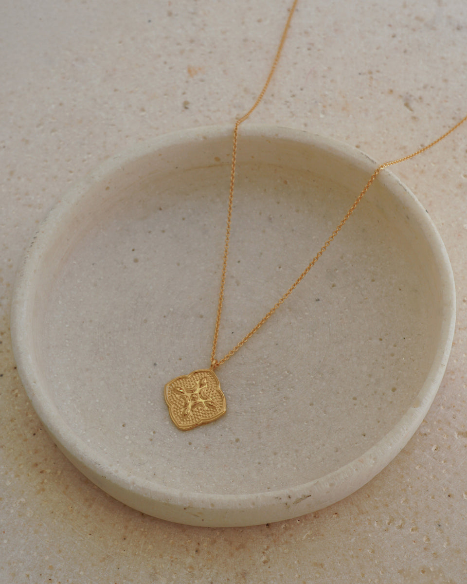 A product shot of the Luna Necklace in gold, featuring a delicate chain and an intricate, quatrefoil-shaped pendant with a textured, embossed design.