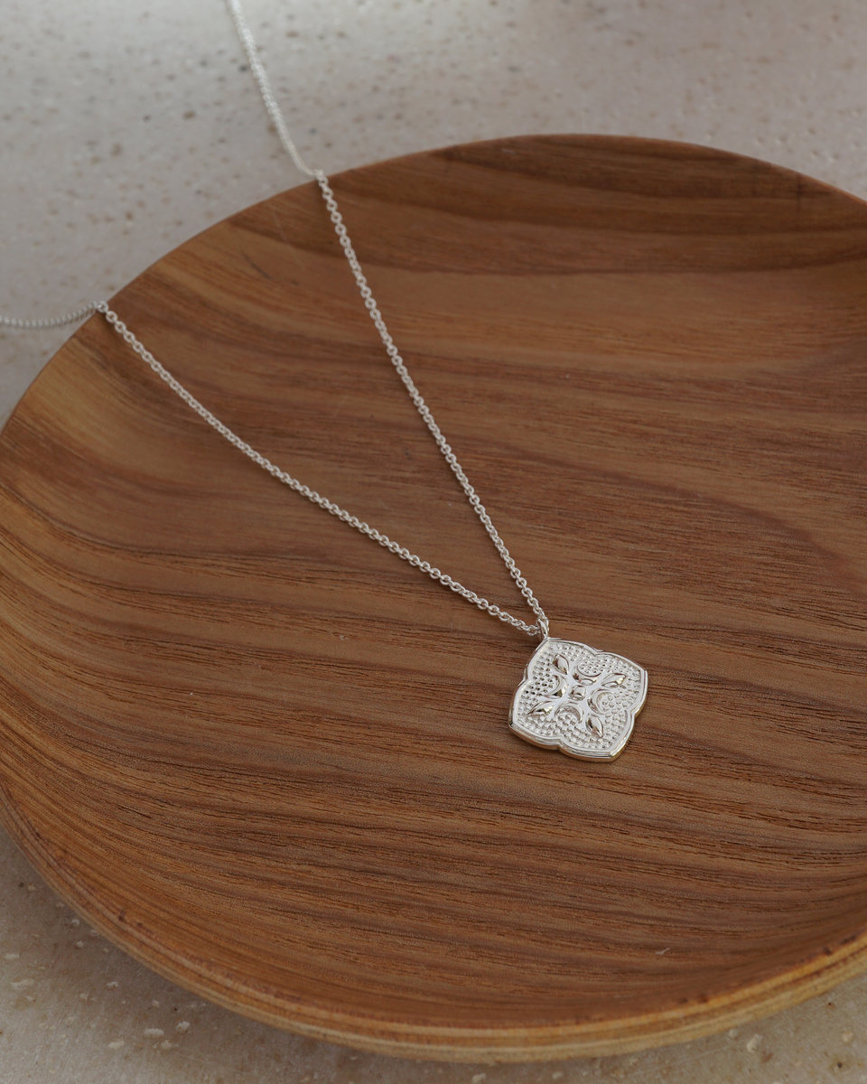 The Luna Necklace in silver resting on a wooden plate, showcasing its delicate chain and a large, intricately patterned, flower-like pendant.