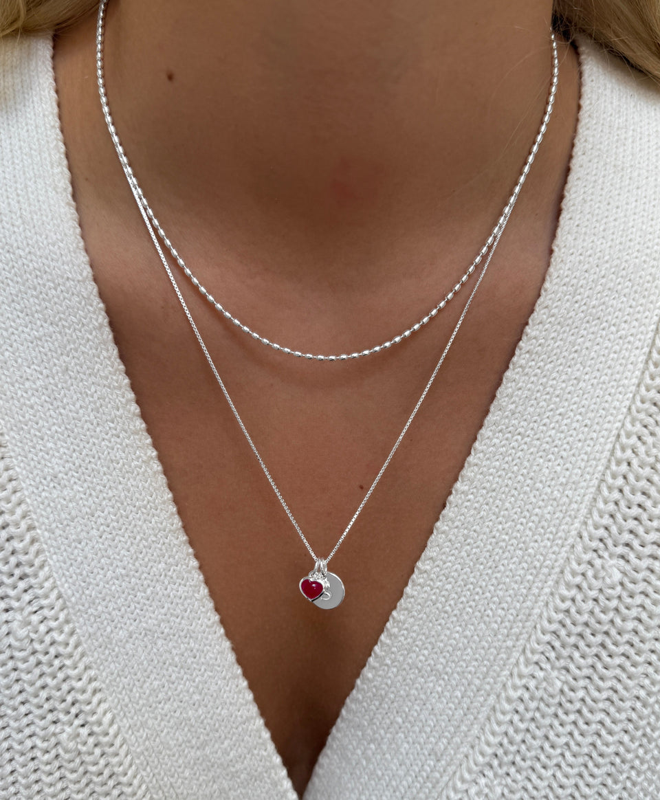 A close-up shot of a model wearing the silver Lunetta Necklace layered with another silver necklace, which features a box chain, a pink quartz heart pendant, and a round letter charm.