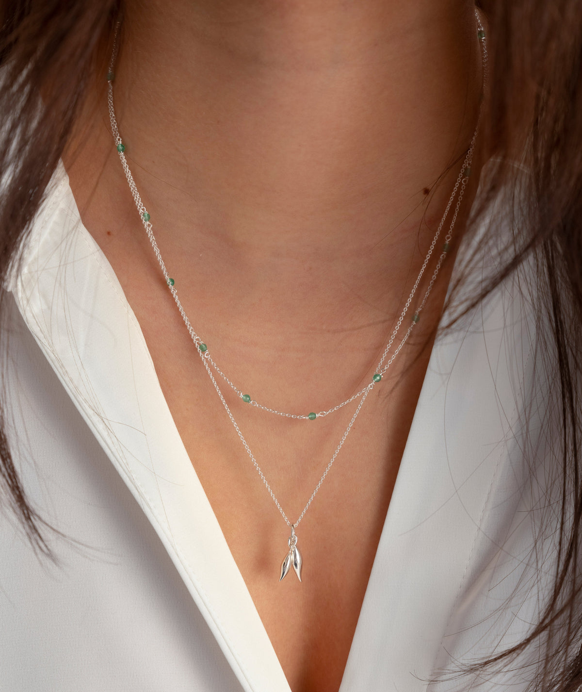 A close-up of a model wearing two layered Syster P necklaces: the Santa Monica Necklace in silver with green onyx stones, and the Melinda Double Petal Necklace in silver with a two-petal pendant.