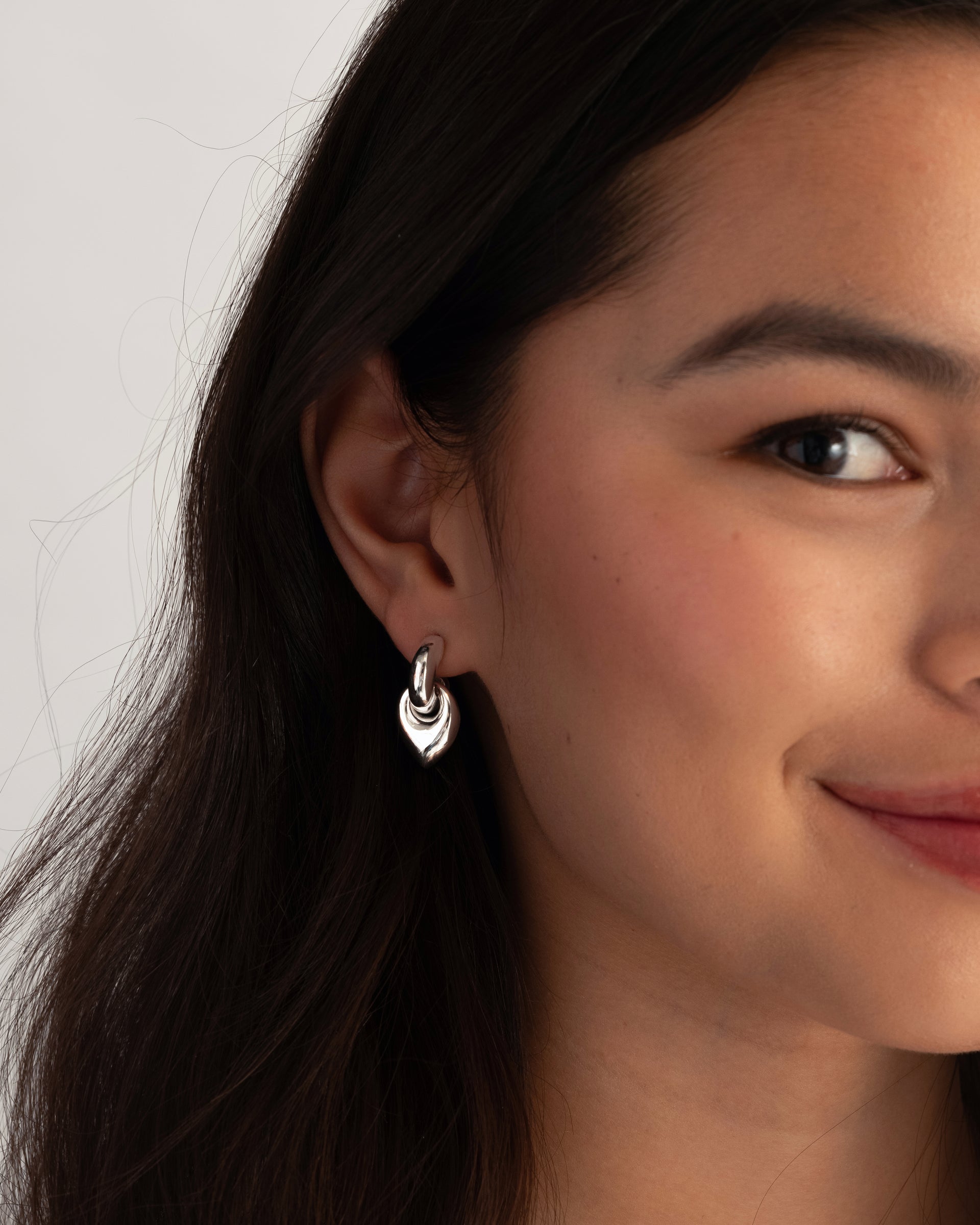 A close-up shot of a model wearing the Melinda Earrings in silver, featuring a chunky hoop with a dangling leaf-shaped pendant.