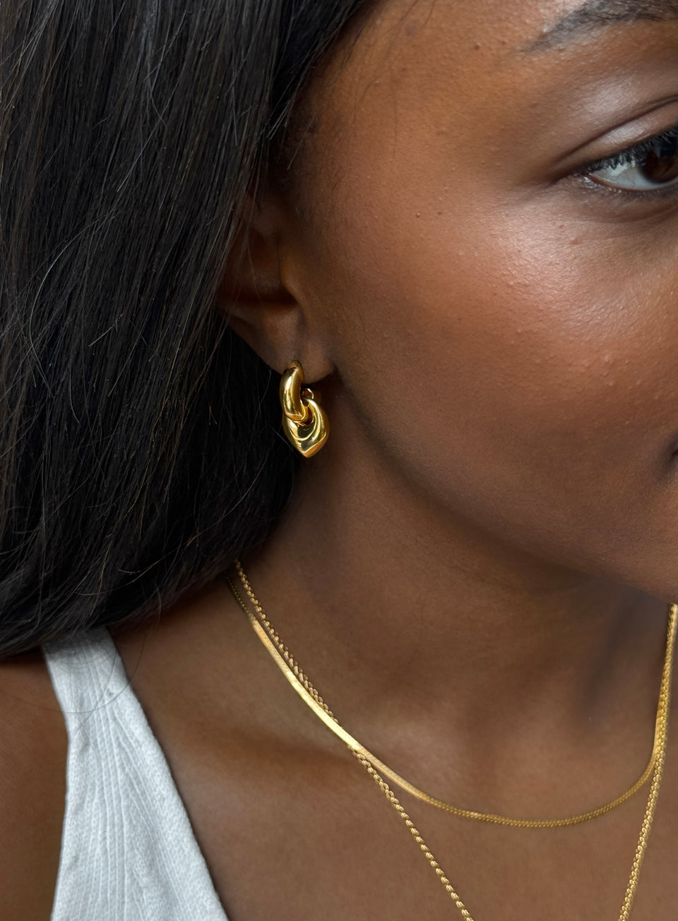 A model wears the Melinda Earrings in gold, featuring a chunky hoop and leaf-shaped pendant, styled with two layered gold necklaces.