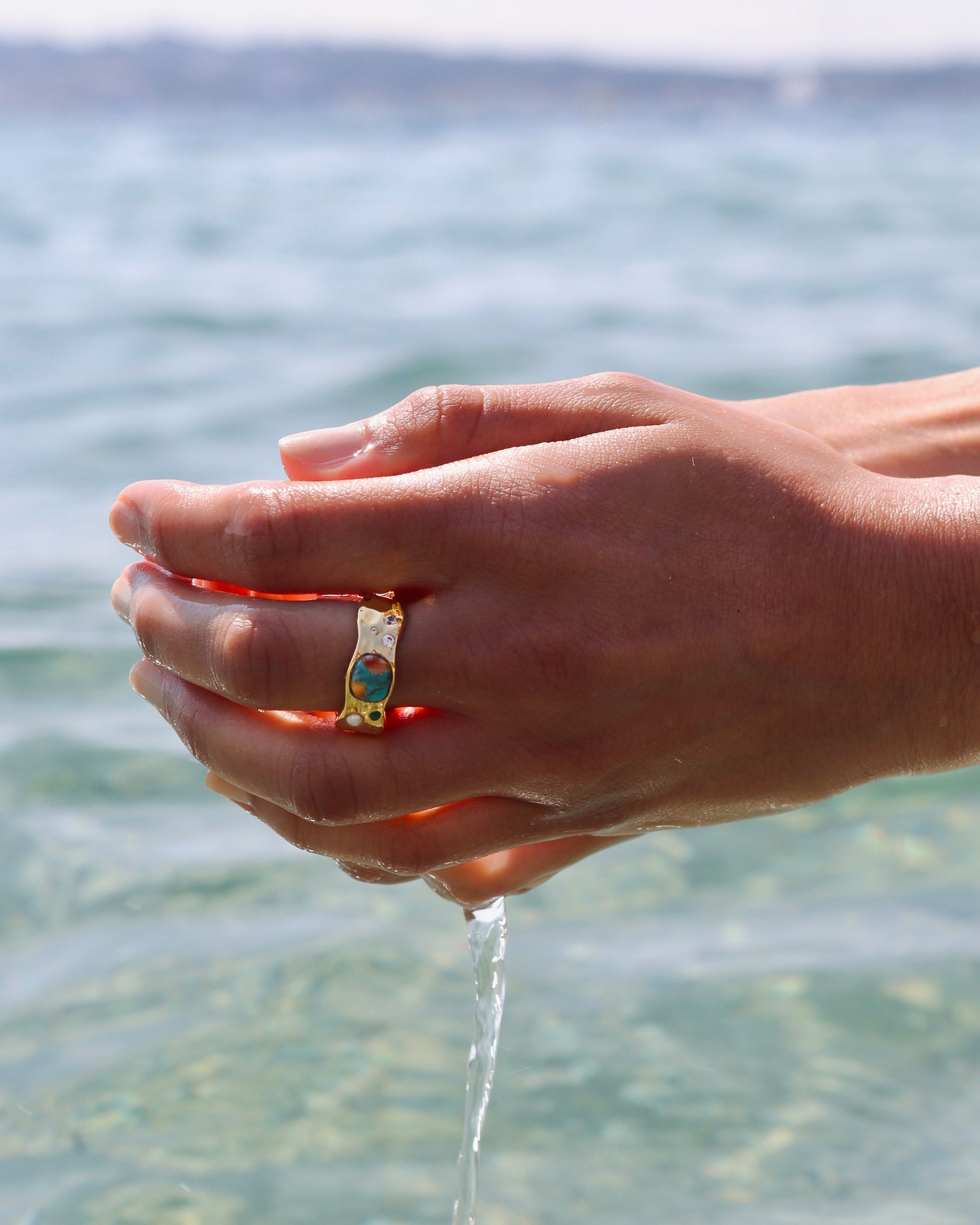 A close-up of a model's hands cupping water from the sea, showcasing the Melting Ring Gold with its unique melted design and colorful gemstone setting.