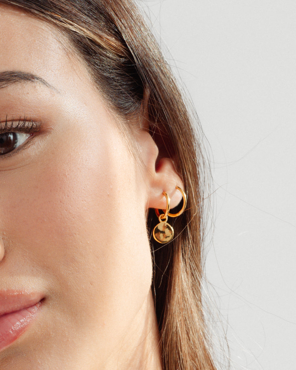 A close-up of a model's ear wearing the gold Minimalistica Hammered Earring with a coin charm and the plain Beloved Fat Medium Hoop.