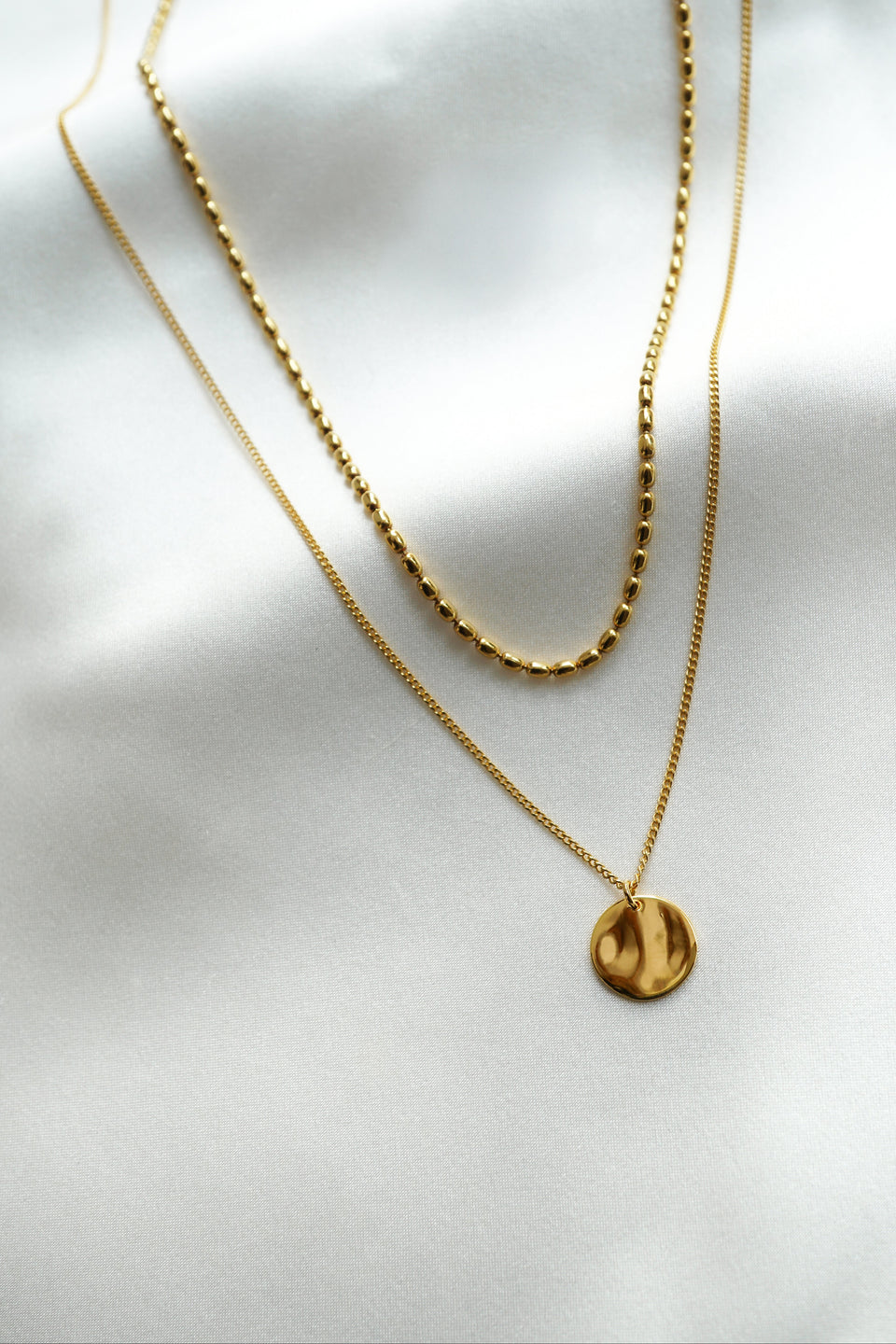 A product shot of the Minimalistica Hammered & Lunetta Necklace Set in gold, layered together. One necklace features a chain of small oval beads, while the other has a fine chain with a round hammered pendant.