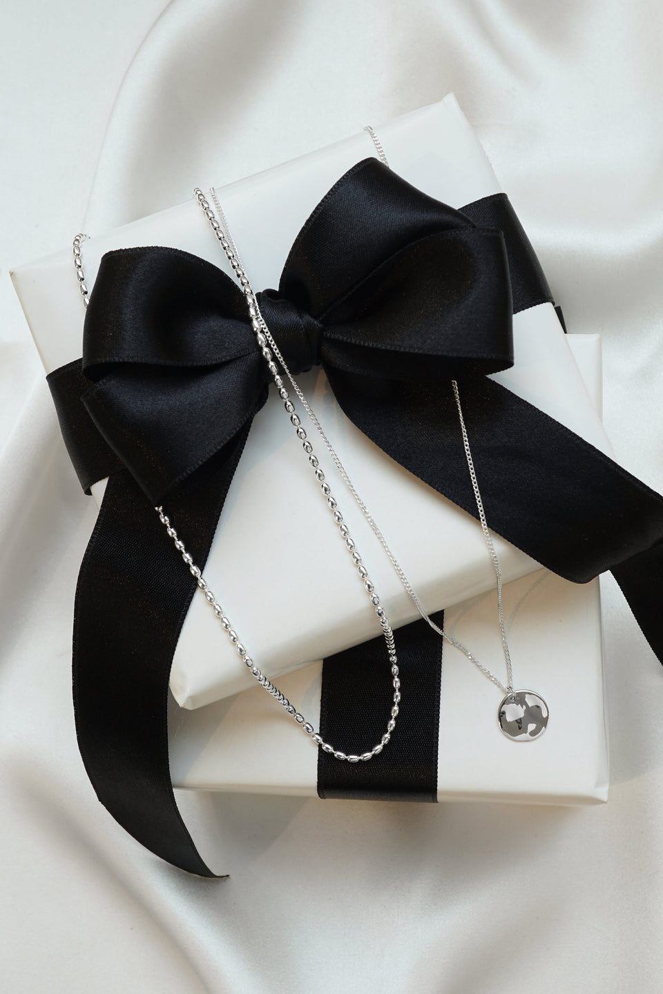 A product shot of the Minimalistica Hammered & Lunetta Necklace Set in silver, featuring the oval-linked Lunetta necklace and the Minimalistica necklace with its round hammered pendant, both displayed on a white gift box with a black bow.
