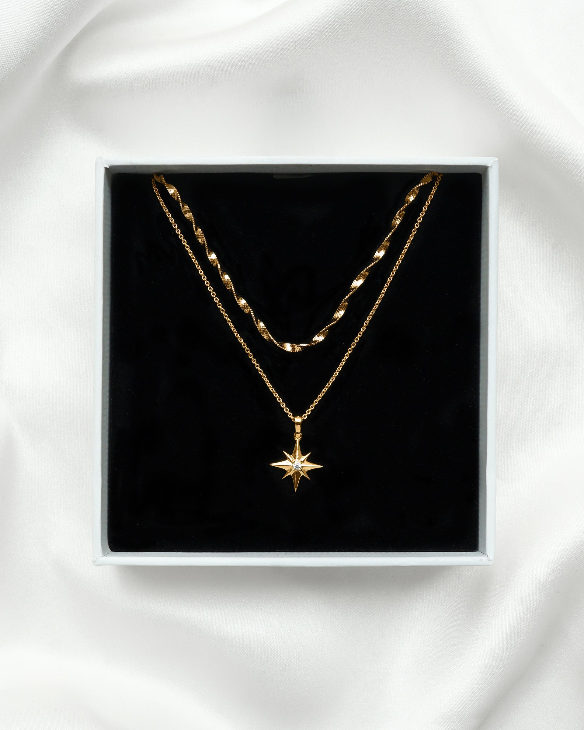 A product shot of the North Star & Herringbone Necklace Set in gold, presented in a Syster P gift box. The set features a twisted herringbone chain layered with a fine chain necklace with a North Star pendant.