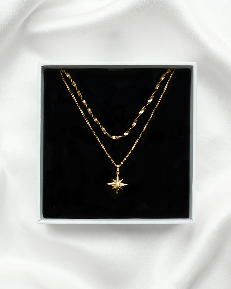 A product shot of the North Star & Herringbone Necklace Set in gold, presented in a Syster P gift box. The set features a twisted herringbone chain layered with a fine chain necklace with a North Star pendant.