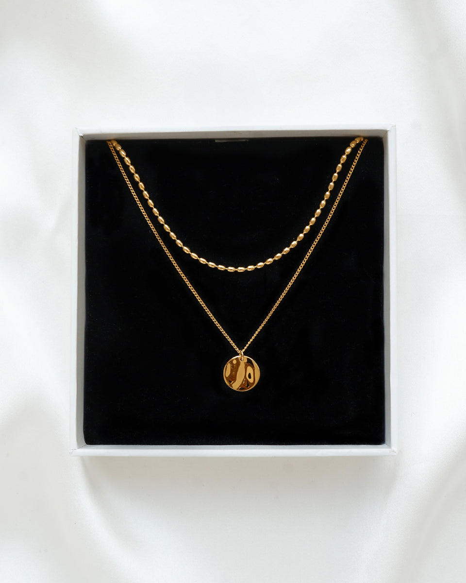A product shot of the Minimalistica Hammered & Lunetta Necklace Set in gold, displayed in a white Syster P jewelry box. The set features two layered necklaces: one with a delicate beaded chain, and another with a fine chain and a round, hammered pendant.