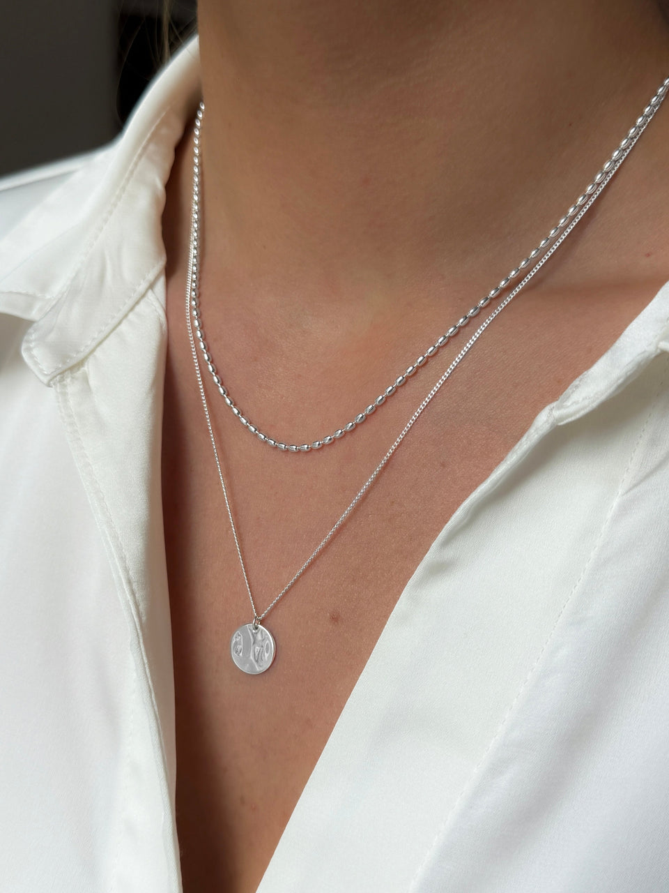 A close-up shot of a model wearing the layered Minimalistica Hammered & Lunetta Necklace Set in silver. The shorter necklace has an oval beaded chain and the longer one has a fine chain with a round, hammered-finish pendant.