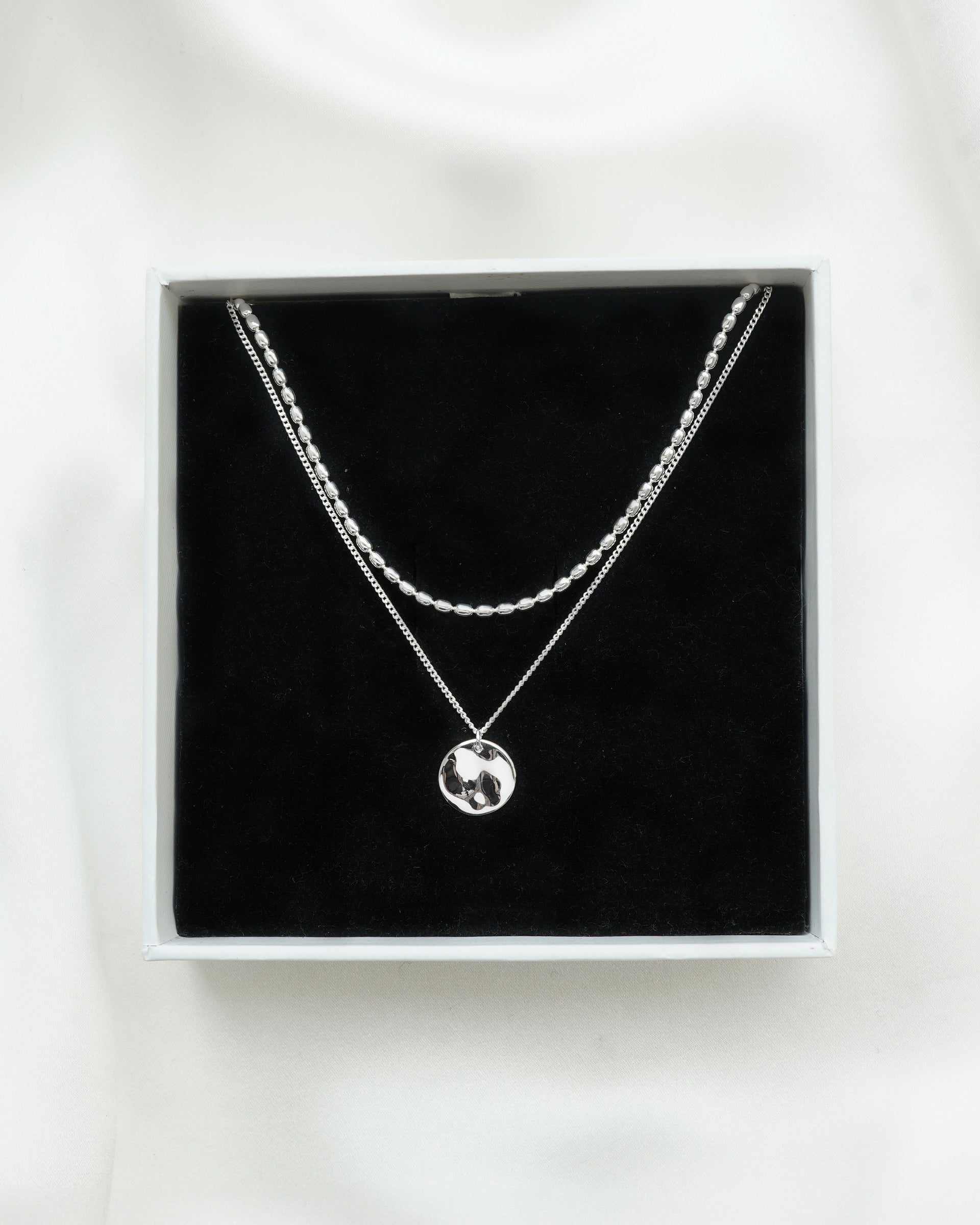A product shot of the Minimalistica Hammered & Lunetta Necklace Set in silver, showcased inside a white Syster P jewelry box. The set features the layered Lunetta necklace with its oval-beaded chain and the Minimalistica necklace with a fine chain and a hammered disc pendant.