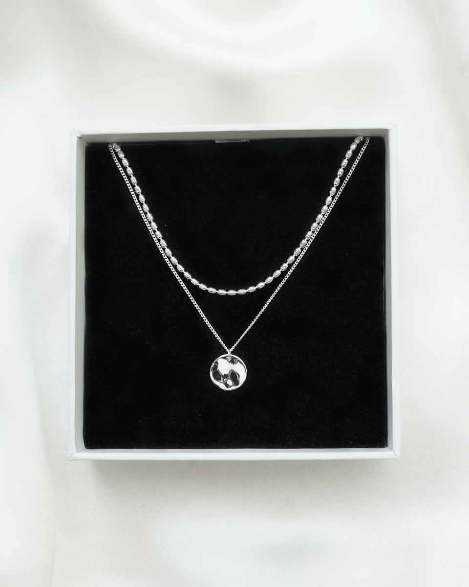 A product shot of the Minimalistica Hammered & Lunetta Necklace Set in silver, showcased inside a white Syster P jewelry box. The set features the layered Lunetta necklace with its oval-beaded chain and the Minimalistica necklace with a fine chain and a hammered disc pendant.