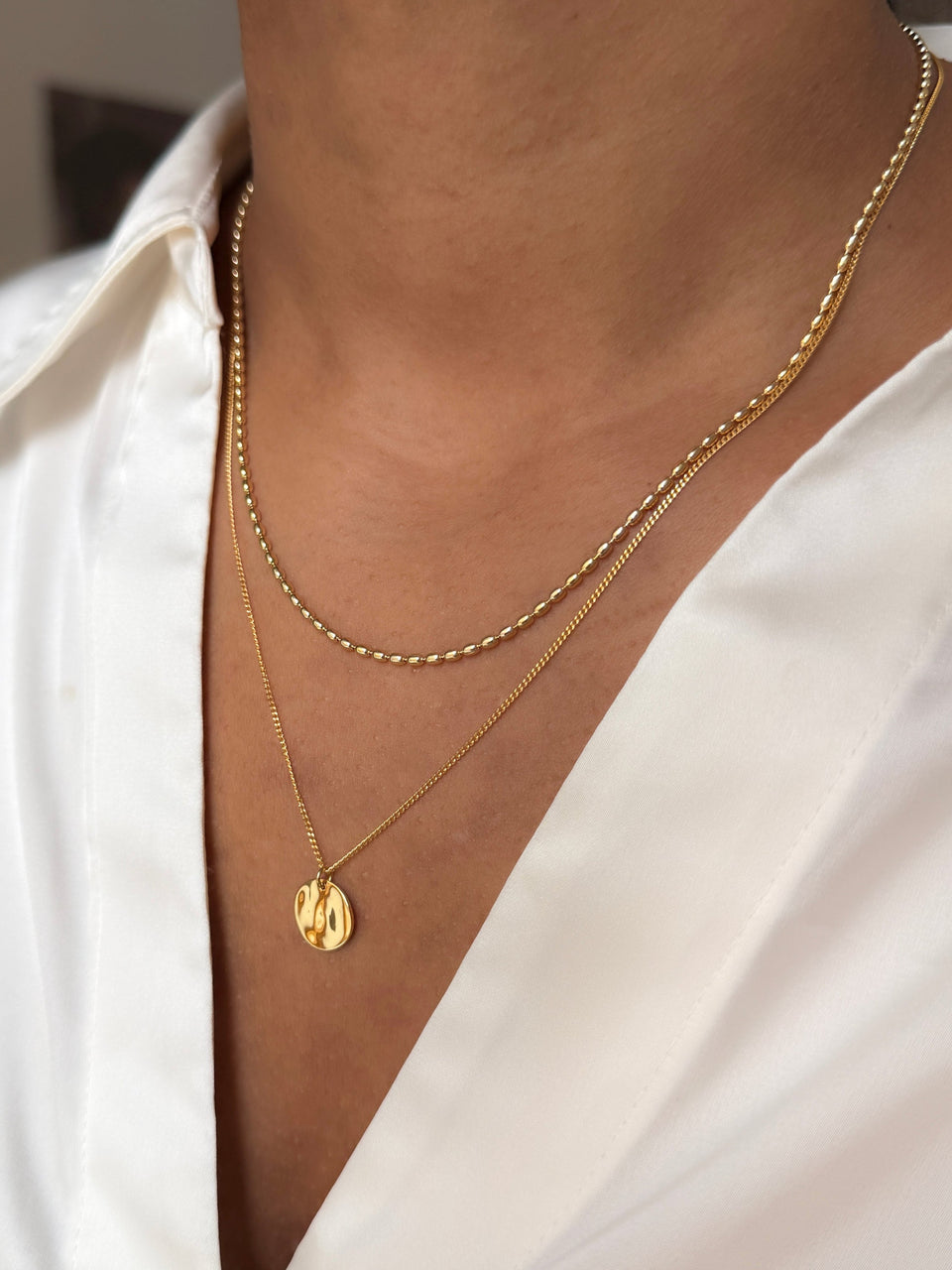 A close-up on a model wearing the Minimalistica Hammered & Lunetta Necklace Set in gold, styled with a white shirt. The image shows the two necklaces layered, one a delicate beaded chain and the other a fine chain with a round, hammered pendant.