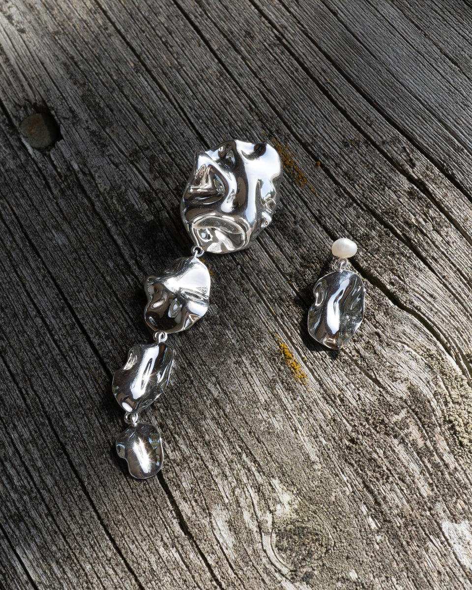 A product shot of the Moulded Long Single Earring and the Moulded Short Single Earring in silver, laid out on a weathered wood background to showcase their organic, molten-like designs.