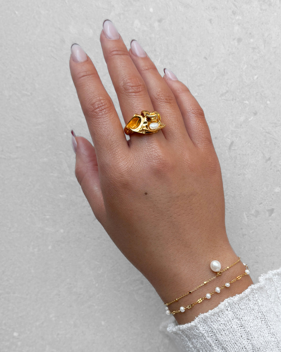 A close-up of a model's hand wearing the Moulded Big Ring in gold, featuring an organic shape and a freshwater pearl. Her wrist is adorned with a stack of gold bracelets, including the Aurelia Bracelet and the Treasure Single Pearl Bracelet, both featuring pearls.