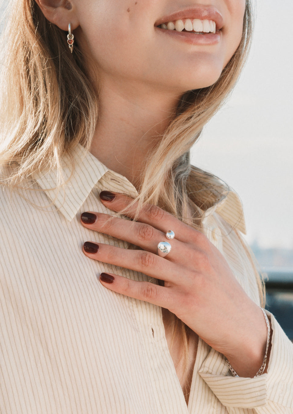 A smiling model showcases a set of silver jewelry, her hand on her chest displaying the bold, drop-shaped Bolded Drops Ring. She also wears a silver hoop earring with a spike and red heart charm, and a silver chain bracelet.