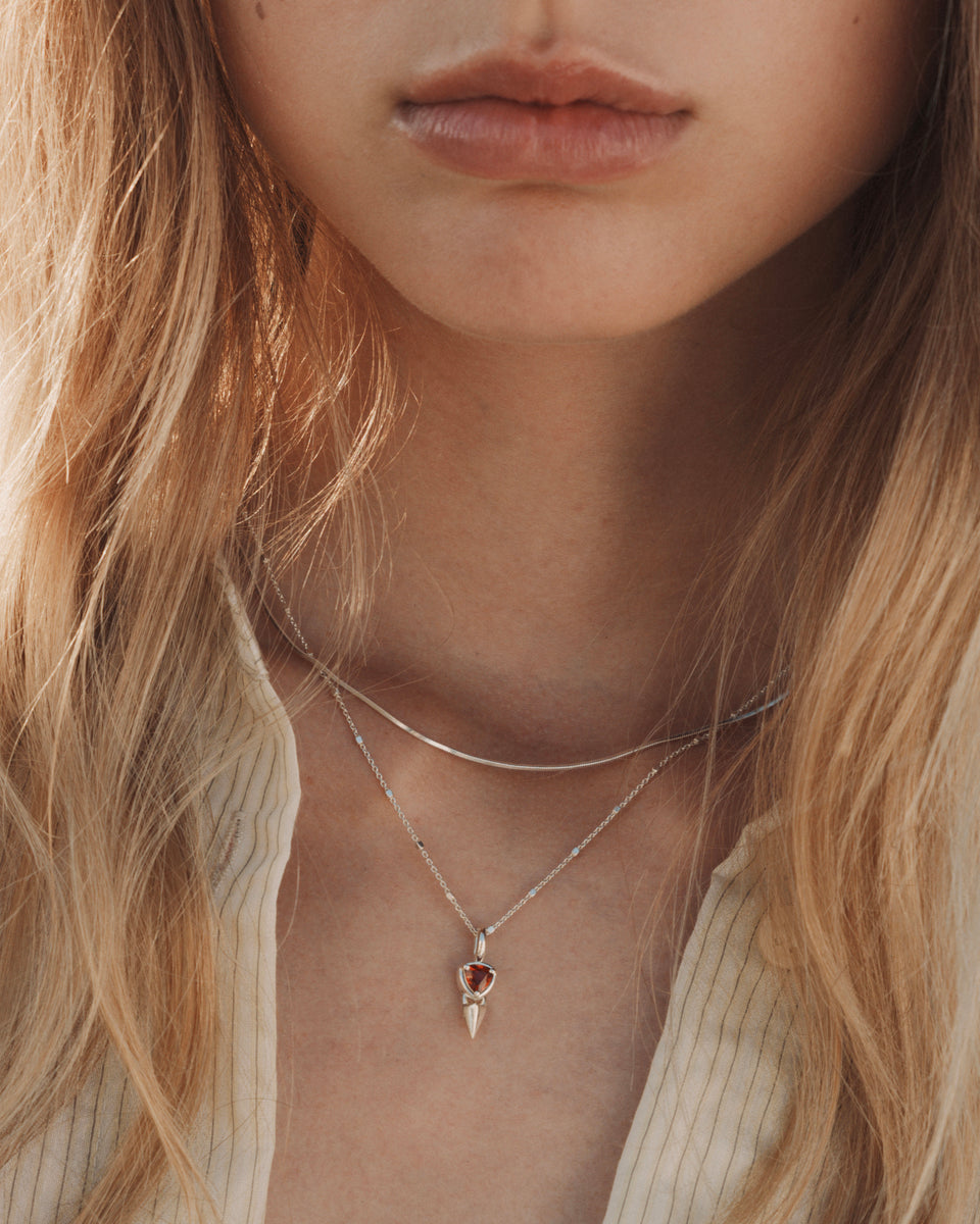A close-up shot of a model wearing the Sheer Mirror Necklace in silver, layered with another silver necklace featuring a spiky pendant with a triangular red stone.
