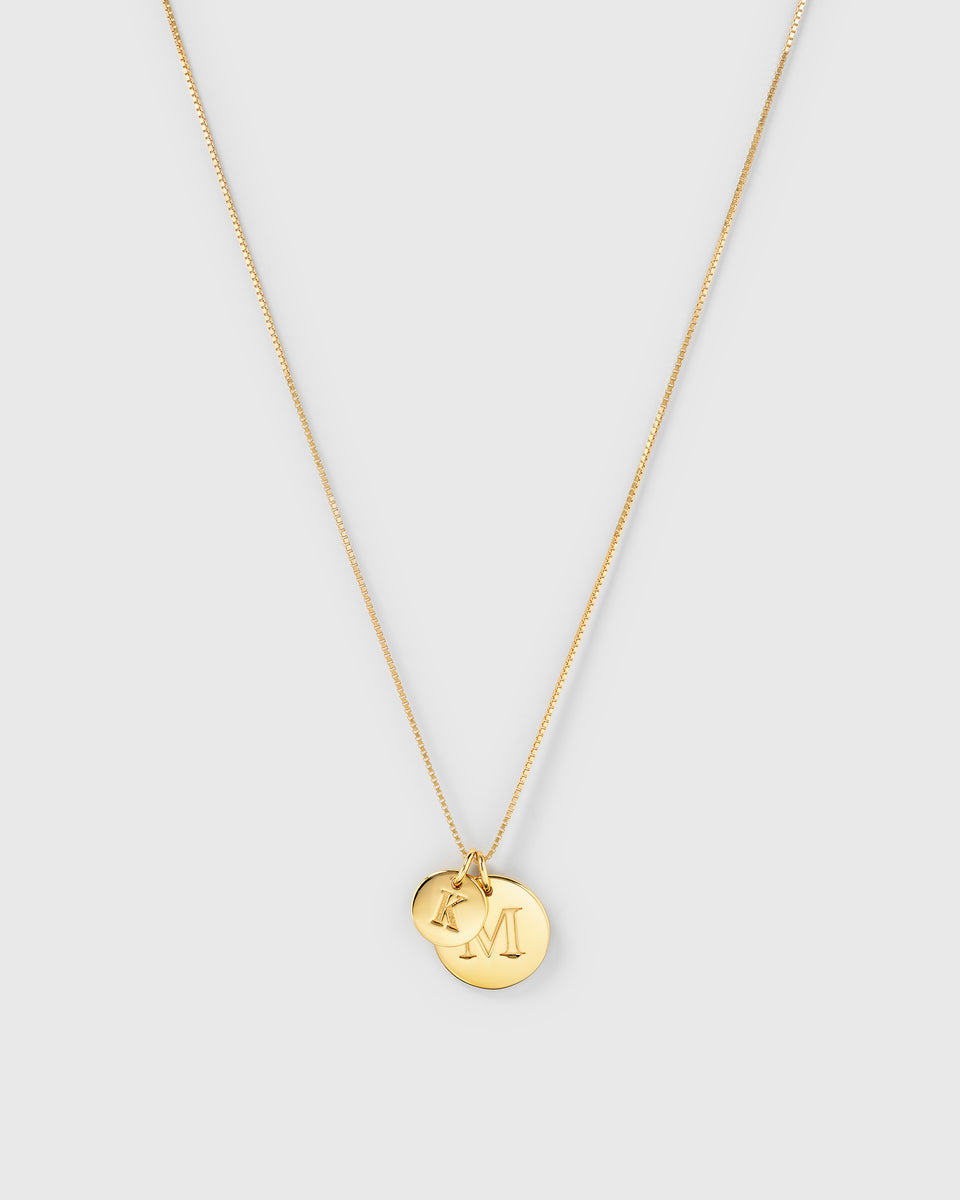 A product shot of a gold necklace styled with a Beloved Big Letter 'M' charm and a Beloved Mini Letter 'K' charm, layered together on a box chain.
