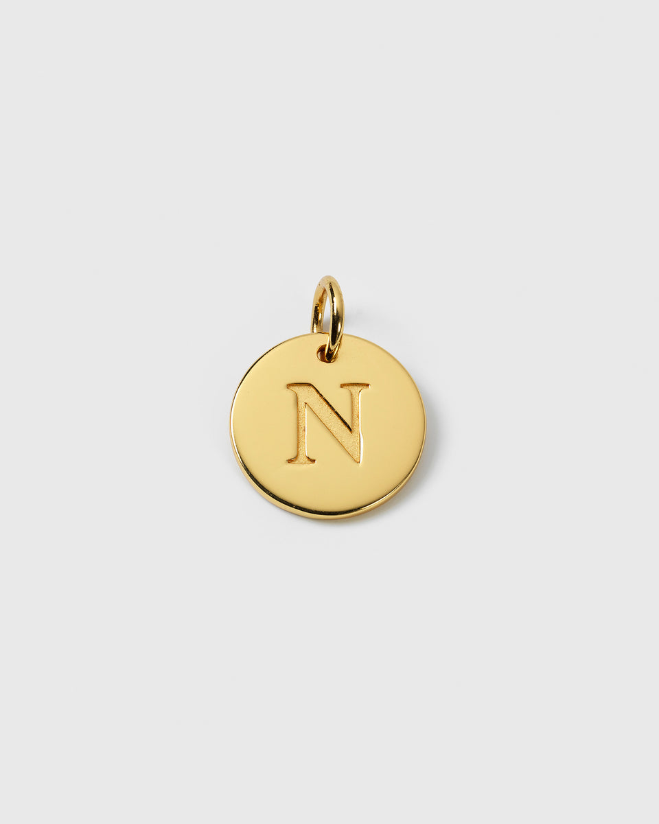 A product shot of the Syster P Beloved Big Letter Gold pendant, showing a circular gold charm with the letter 'N' engraved in the center.