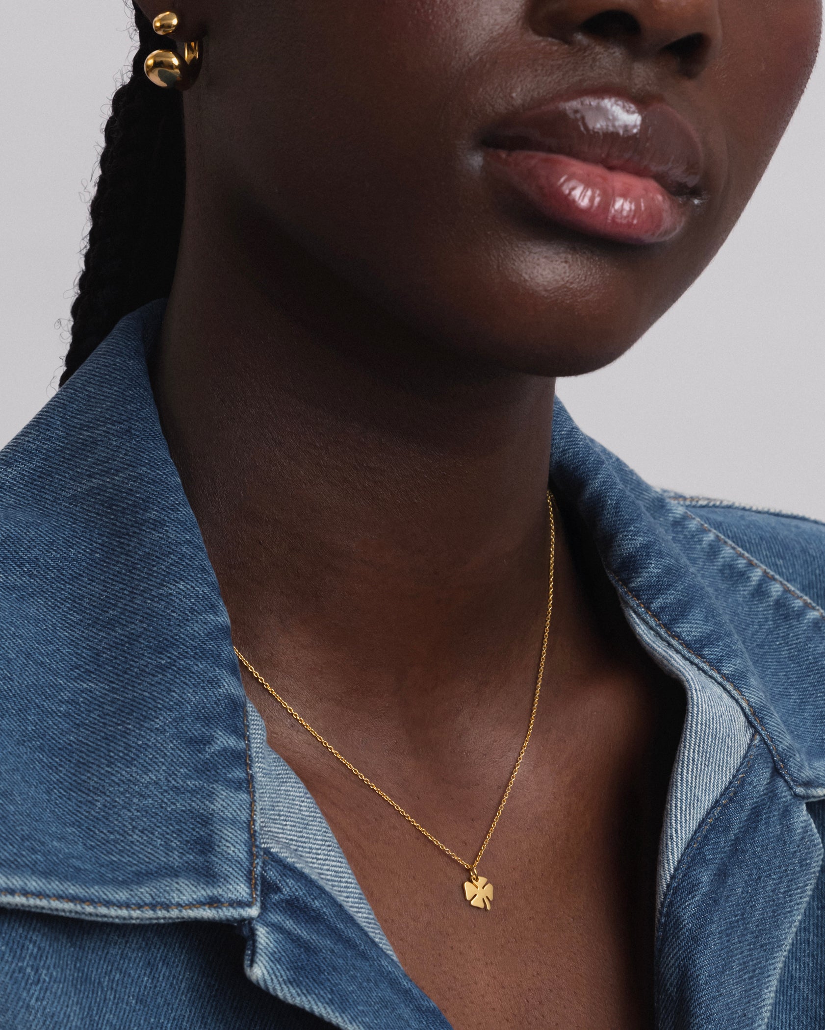 A close-up shot of a model wearing the Bring Me Luck Necklace in gold, which features a fine chain and a small four-leaf clover pendant, paired with chunky gold hoop earrings.