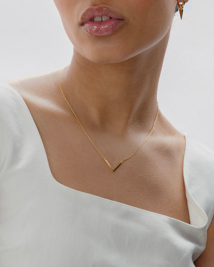 A close-up of a model wearing the Strict V Necklace Gold, featuring a V-shaped pendant on a fine chain, paired with a small gold hoop earring with a spike charm.