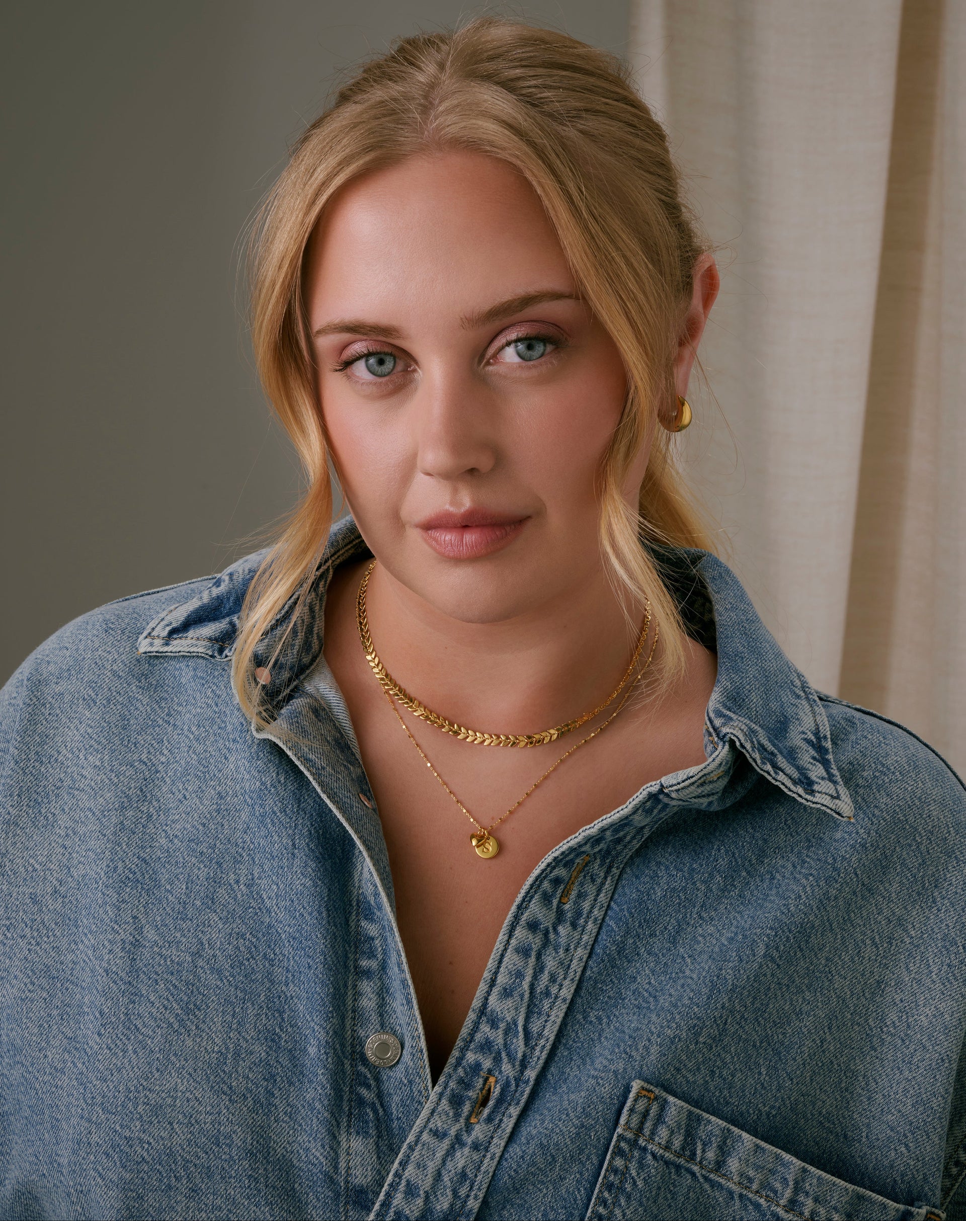 A model wearing a gold necklace with a leaf-like pattern, layered with a delicate gold chain with charms