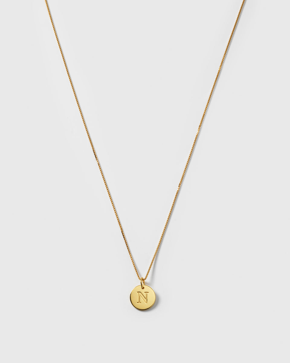 A product shot of the Syster P Beloved Long Box Chain in gold, styled with a small, circular gold pendant featuring an engraved letter 'N'.