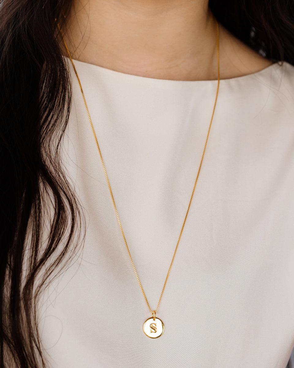 A model wears the Beloved Long Box Chain Gold, styled with a round gold pendant featuring the initial 'S'.