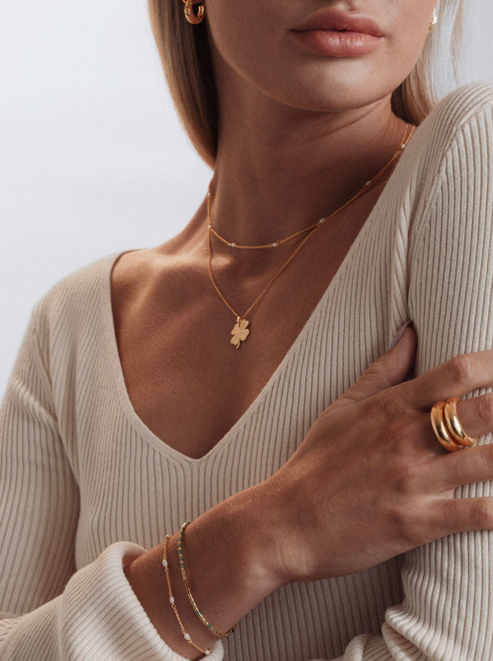 A model wearing the Beaches Fortune Necklace Gold with its clover pendant, layered with a delicate pearl necklace. The look is styled with small gold hoops, a chunky gold ring, and a set of fine bracelets.