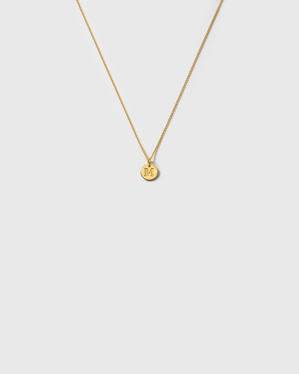 A product shot of the Beloved Baby Chain Gold, a fine anchor chain necklace, styled with a small, round gold pendant engraved with the initial 'M'.