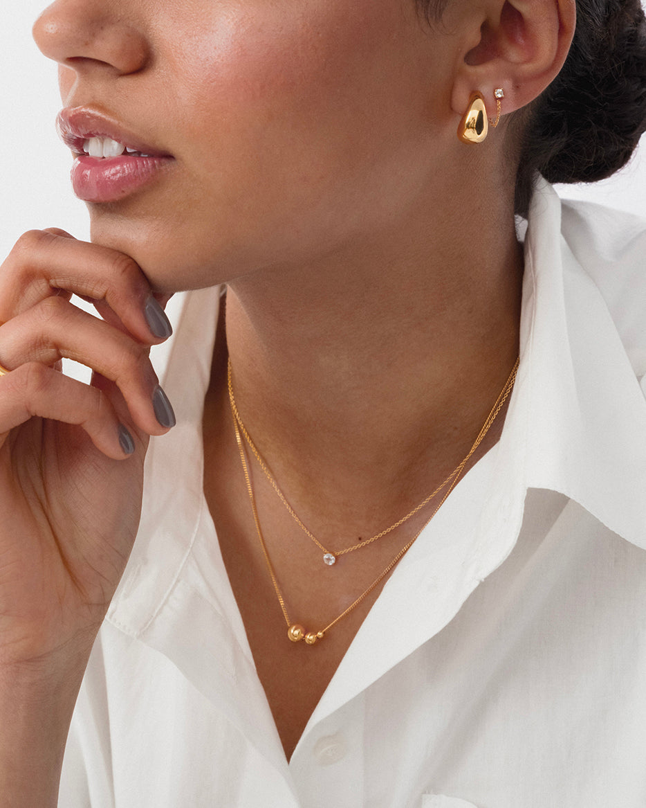 A close-up of a model wearing the Universe Necklace Gold, featuring three gold balls on a fine chain. She has layered it with a second gold necklace with a single stone, and is also wearing a chunky gold hoop and a small stud earring.