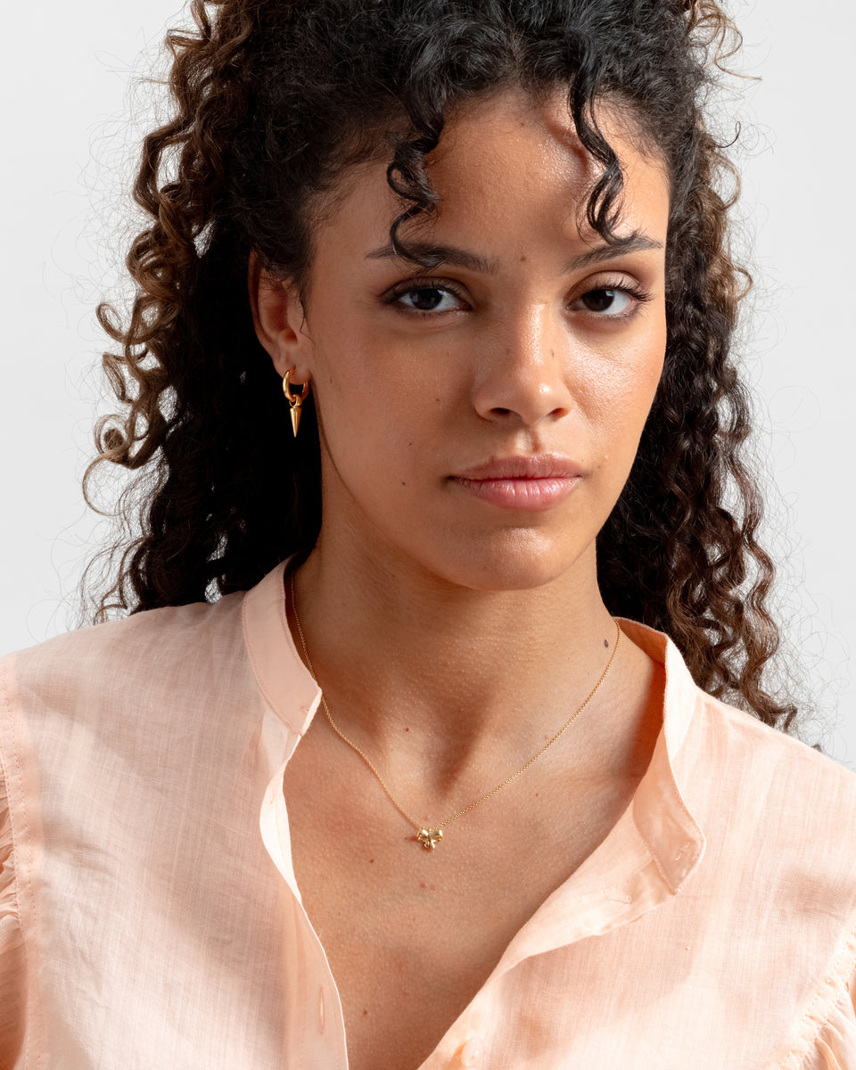 A close-up shot of a model wearing the Rosie Mini Necklace in gold, featuring a delicate chain and a small bow pendant, along with a gold hoop earring with a spike charm.