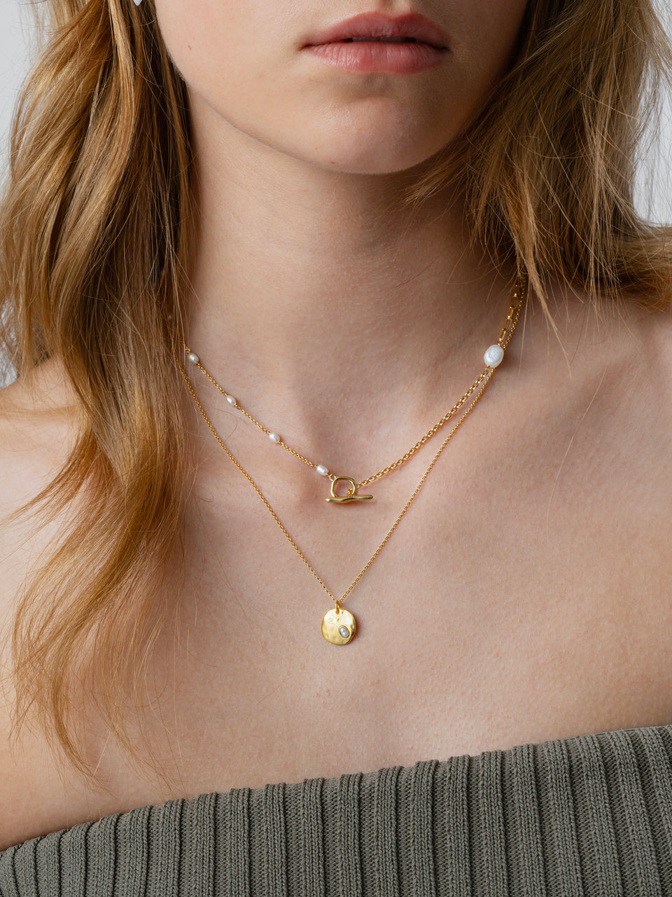 A close-up of a model's neck, showcasing the gold Paxton Short Necklace with its toggle clasp and small pearls, layered with a second longer gold necklace featuring a hammered pendant.