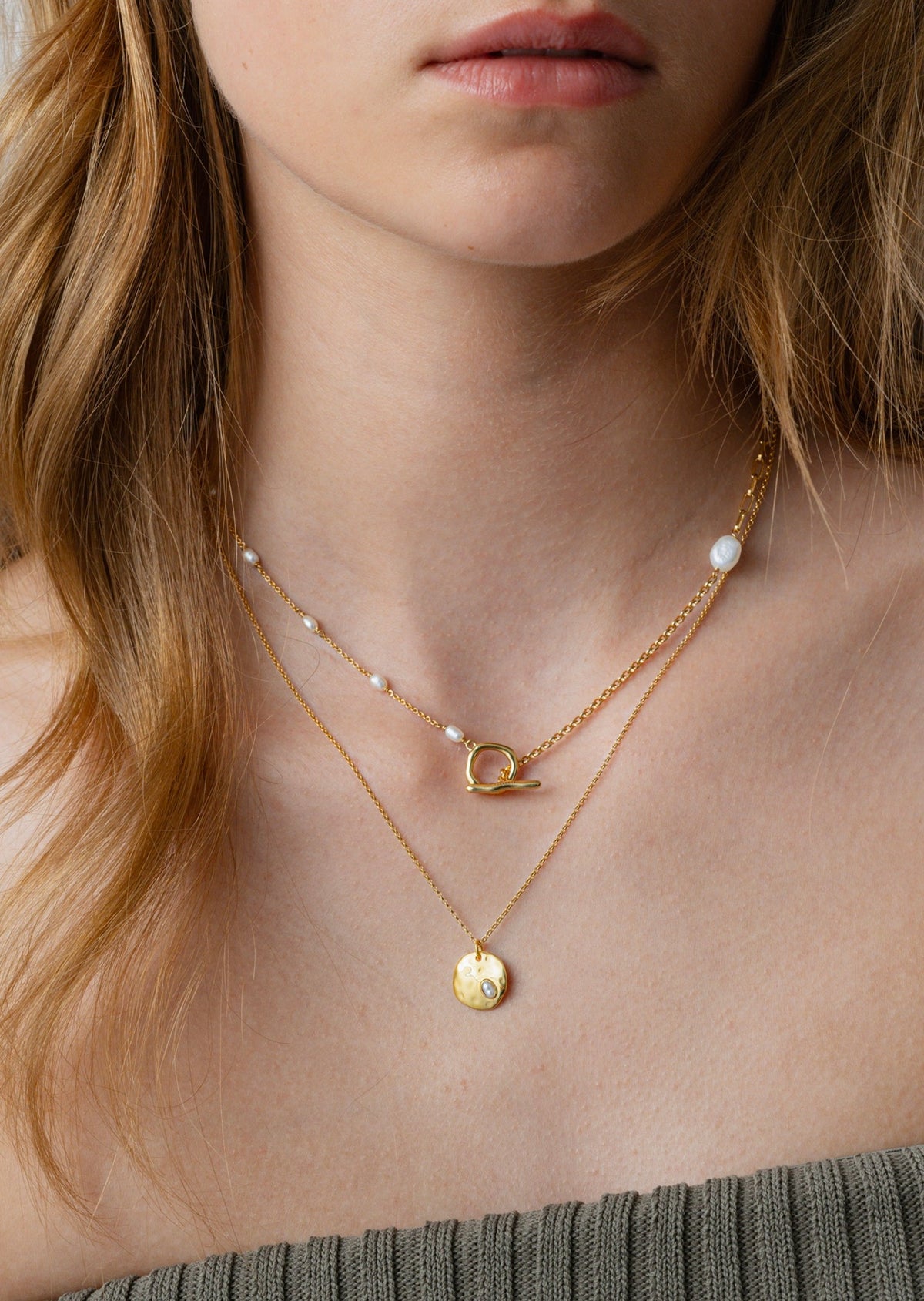 Close-up on a model wearing the Paxton Necklace Gold with its hammered pearl pendant, layered with a shorter gold chain necklace featuring small pearls and a toggle clasp.