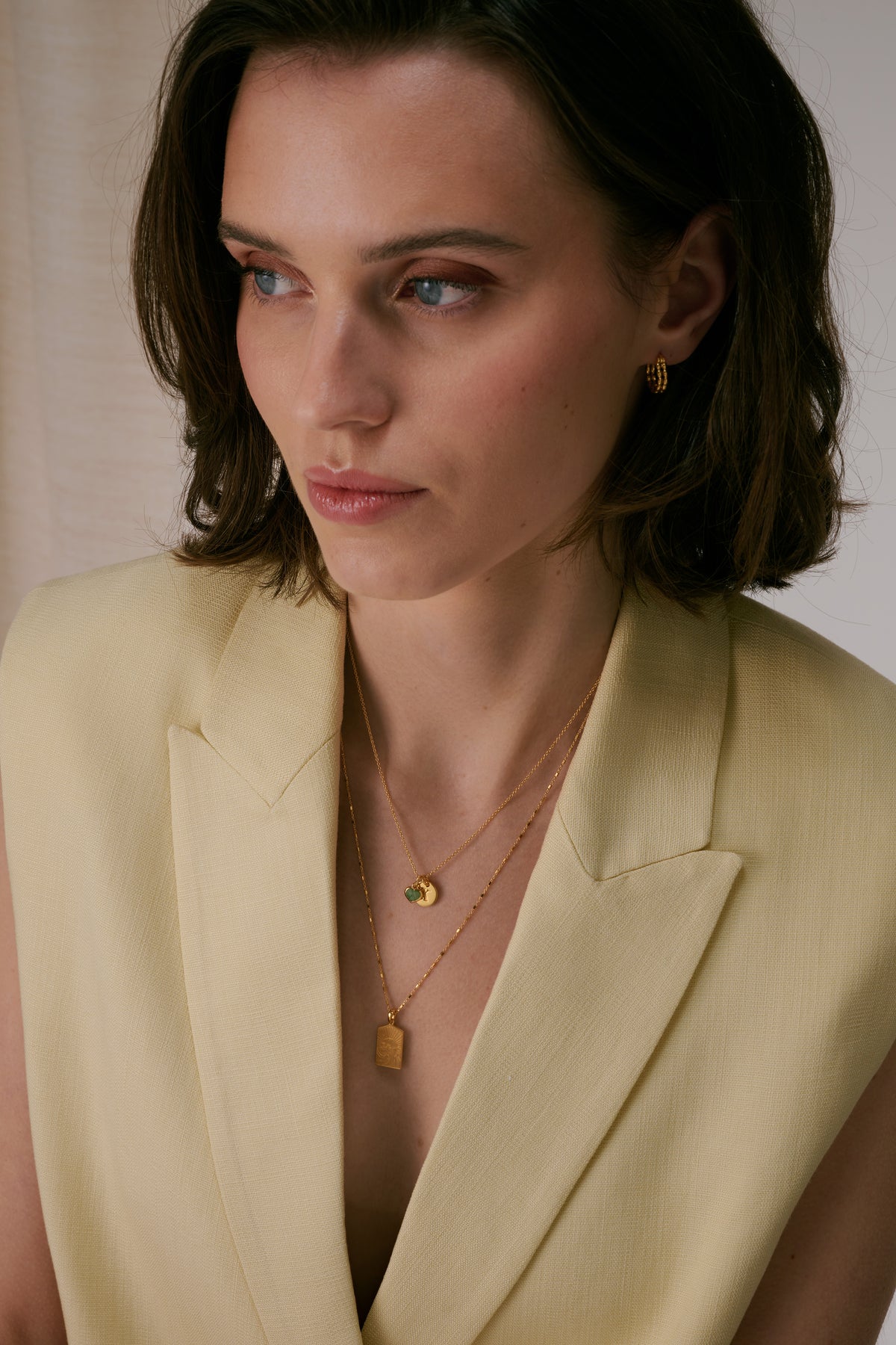 Two necklaces paired with textured gold hoop earrings. The shorter necklace features a green aventurine heart pendant alongside a letter pendant, while the longer chain is adorned with a rectangular gold pendant.
