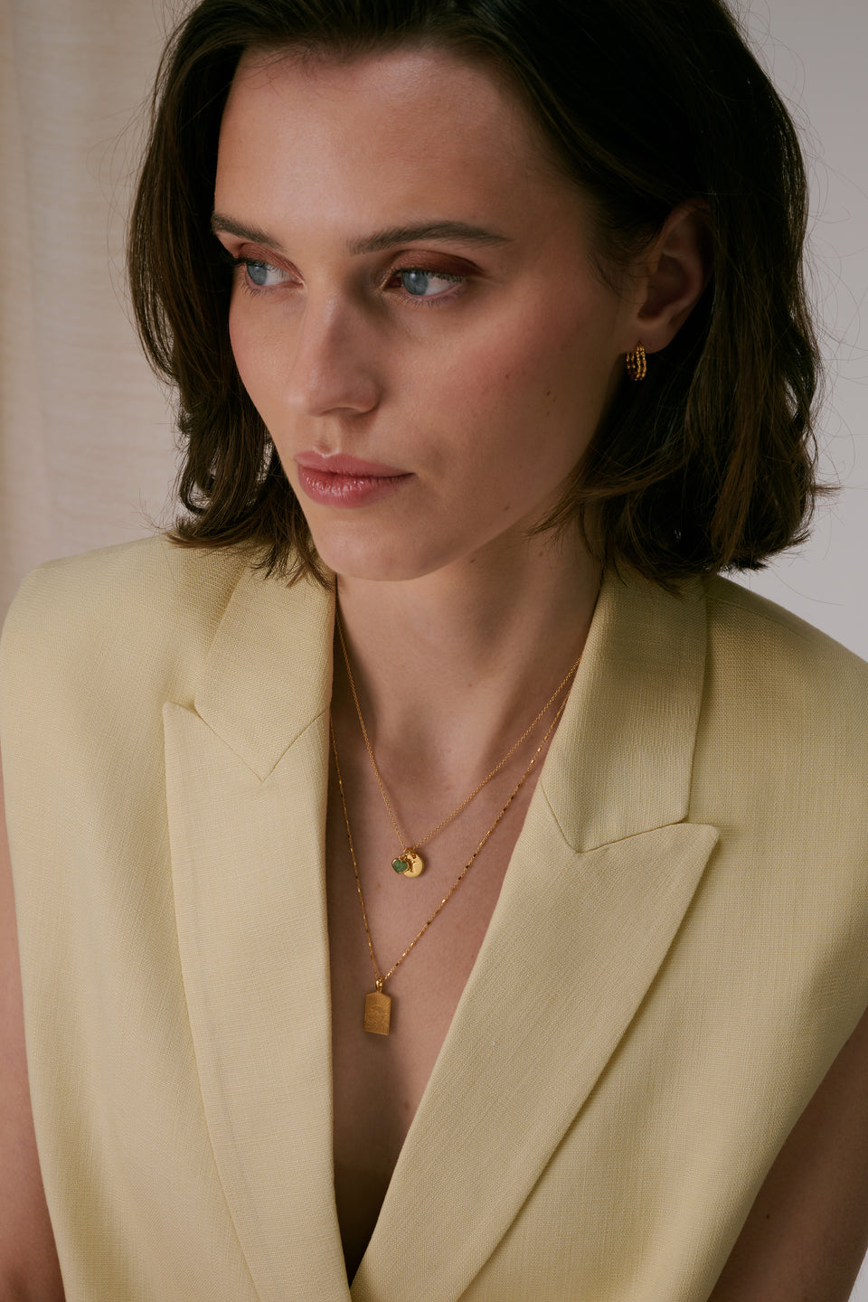 Two necklaces paired with textured gold hoop earrings. The shorter necklace features a green aventurine heart pendant alongside a letter pendant, while the longer chain is adorned with a rectangular gold pendant.