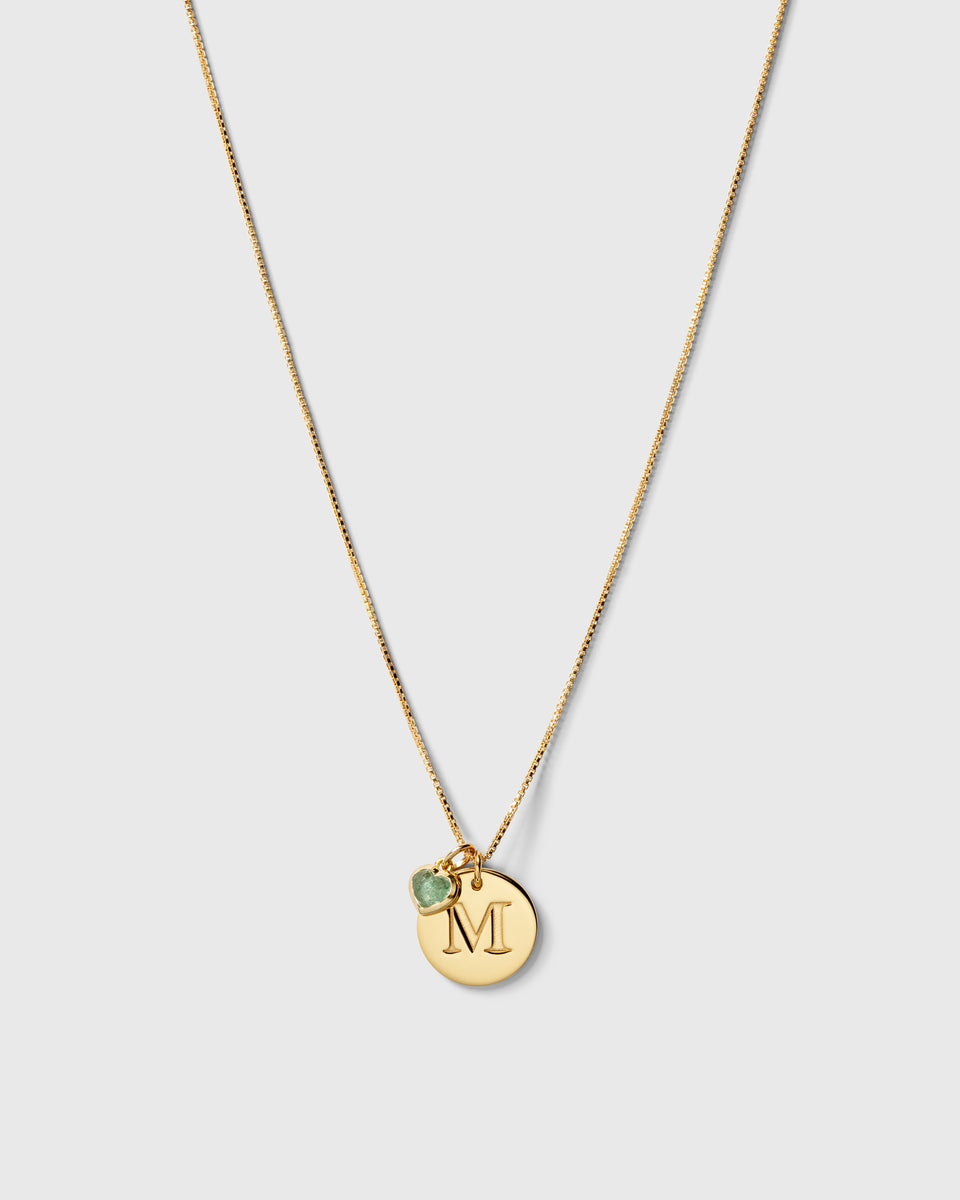A product shot of the Beloved Heart Pendant in gold with a green aventurine stone, styled on a fine gold chain with a round gold pendant engraved with the letter 'M'.