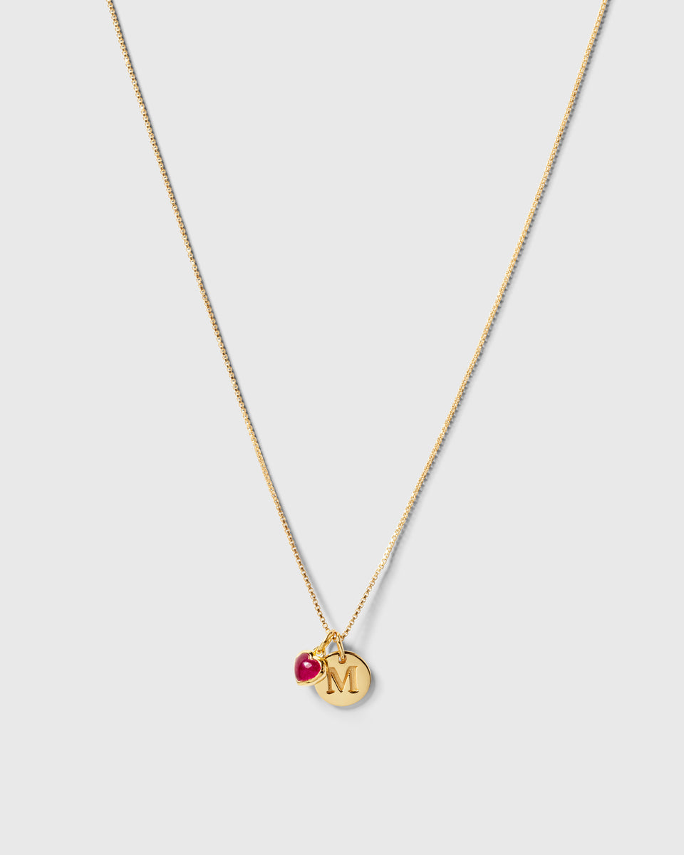 A product shot of a gold necklace featuring the Beloved Heart Pendant with a pink quartz stone, styled alongside a round gold pendant with the initial 'M'.