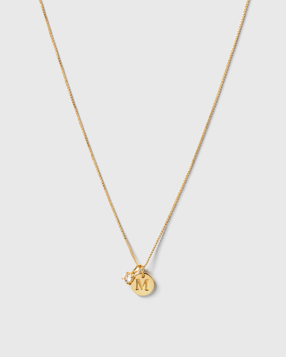A product shot of a gold necklace featuring the Beloved Solo Nova Pendant with its white topaz stone, styled alongside a round gold pendant engraved with the letter 'M'.