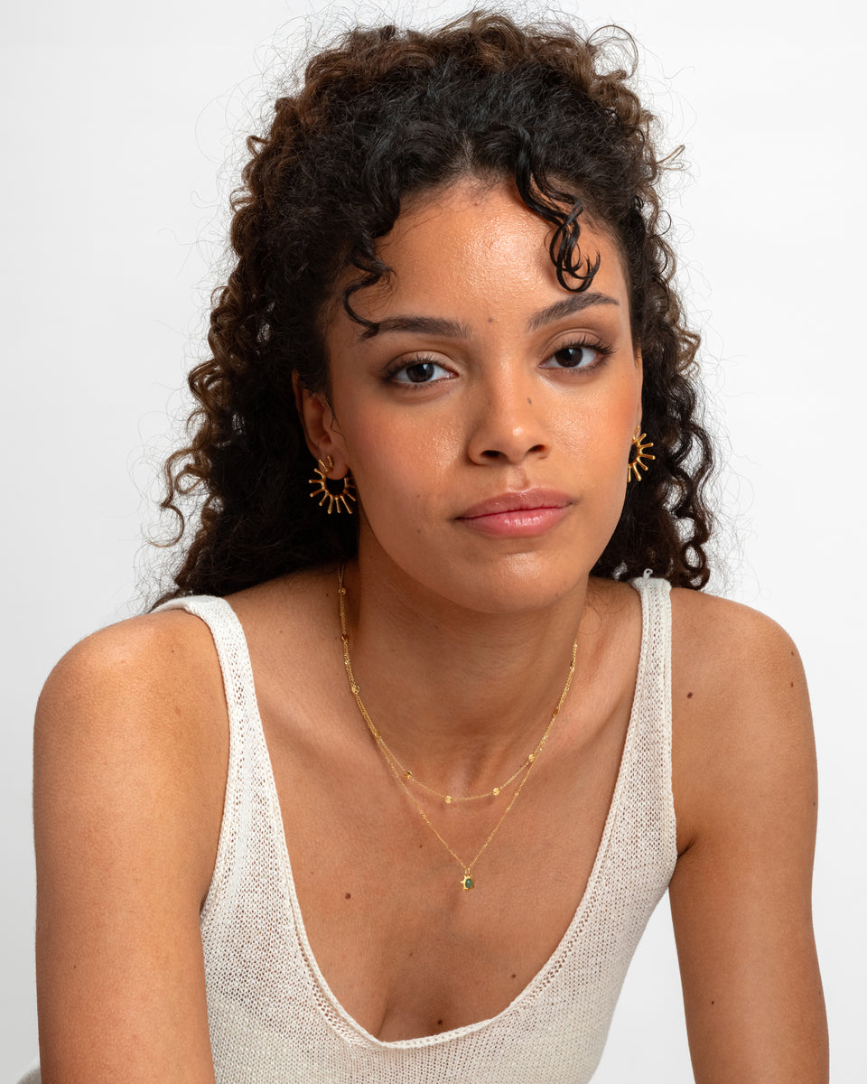 A model wearing the Florentina Necklace Gold with its green jade flower pendant, layered with another delicate gold chain necklace. She also wears a pair of gold sunburst-style hoop earrings.