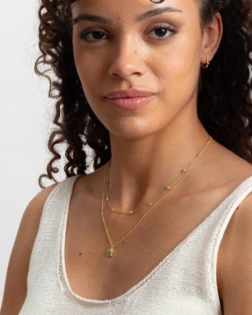 A close-up shot of a model wearing the Florentina Necklace in gold with a green jade flower pendant, layered with a shorter beaded gold chain necklace and paired with a small gold hoop earring.