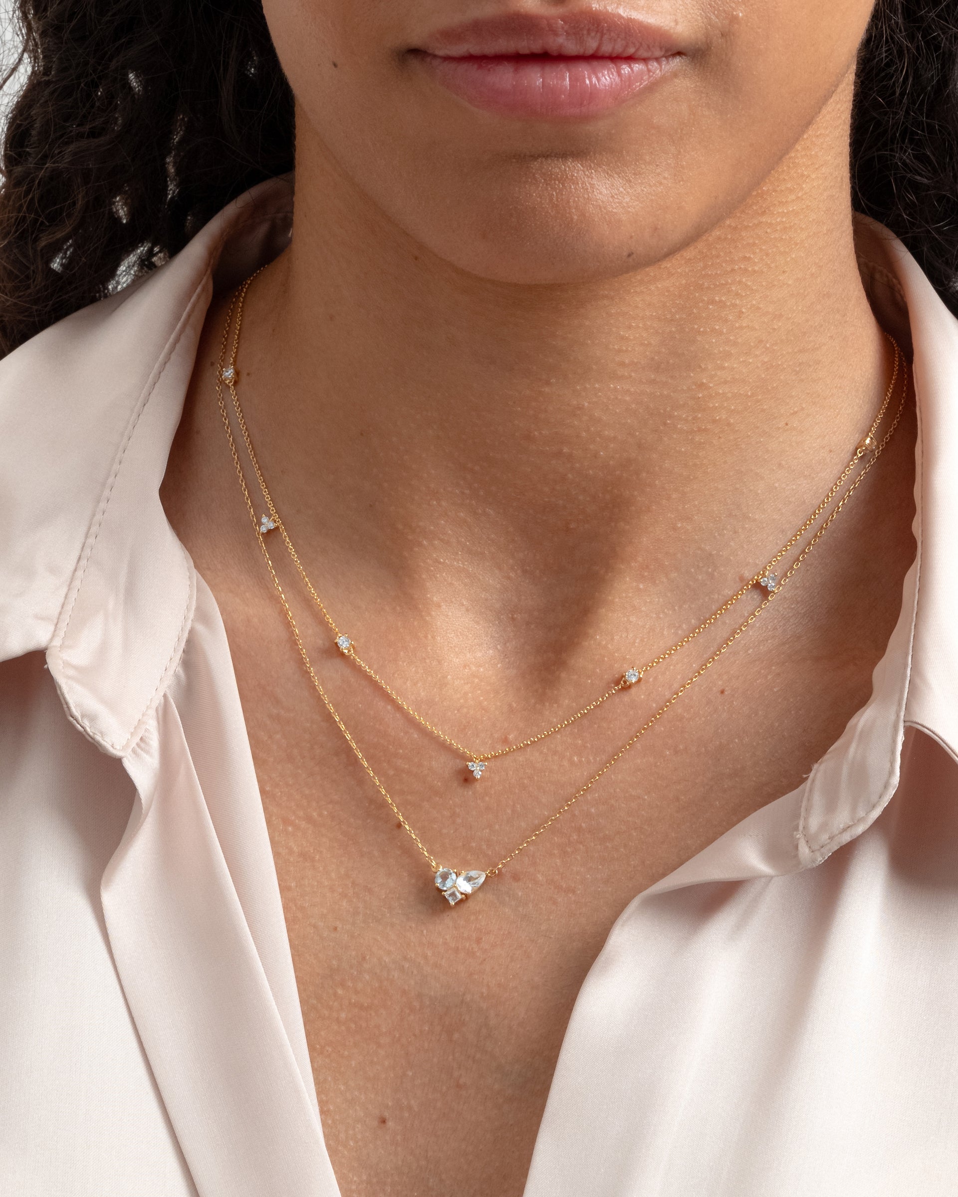 A close-up shot of a model wearing the Celestia Necklace in gold, a fine chain adorned with small, spaced-out cubic zirconia stones, layered with another gold necklace featuring a cluster stone pendant.