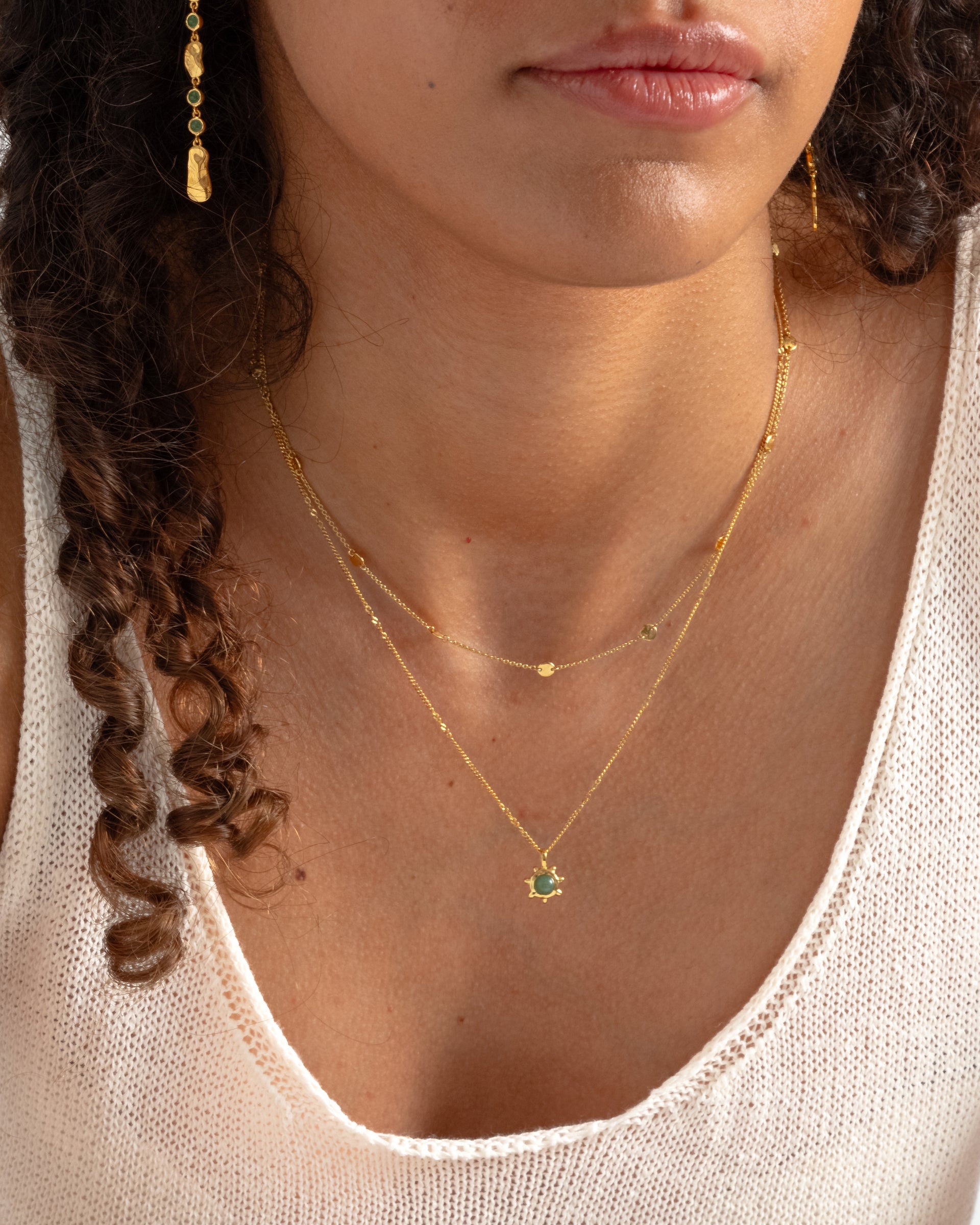 A model wears a layered look with the Solea Necklace in gold, which features a fine chain with small hammered discs, worn with a longer necklace with a green sun pendant and matching gold drop earrings.
