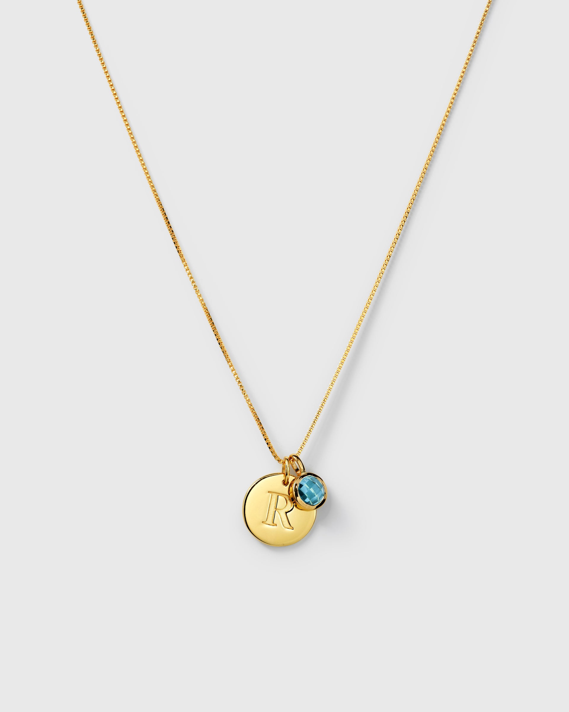 Gold necklace featuring a September birthstone blue topaz pendant and a round letter charm on the same chain.